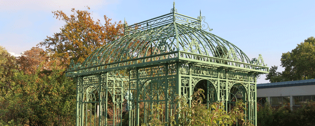 Das Bild zeigt die grüne Metallkonstruktion des Rosenpavillons vor hellblauem Himmel und dem herbstlichen Laub der umgebenden Sträucher und Bäume.